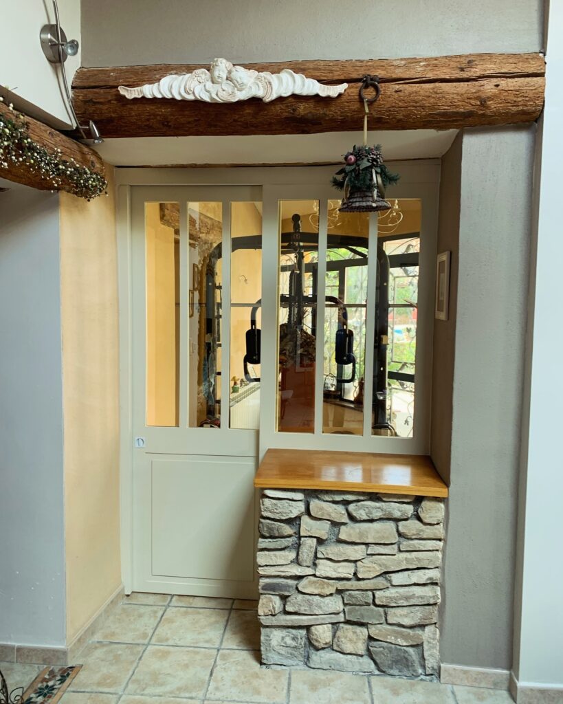 Décoration rustique : porte, bois, pierre et chérubins Porte vitrée avec cadre clair et décorations rustiques : poutre en bois, chérubins sculptés et carillon suspendu au-dessus d'un banc en pierre.