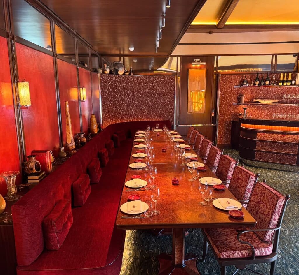 Longue table dressée pour dîner dans un restaurant au décor Art Déco luxueux, dominé par des teintes rouge velours et bois sombre.