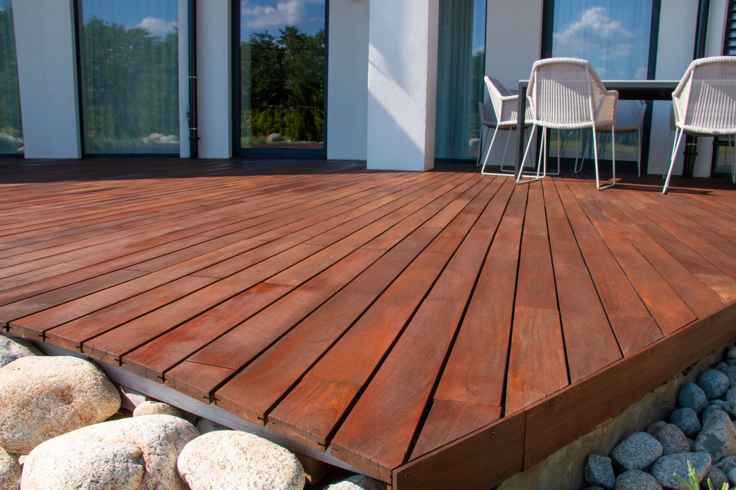 Terrasse en bois rouge foncé, près d'une façade blanche avec grandes baies vitrées reflétant le ciel et des arbres. Gros plan sur les galets clairs et gris.