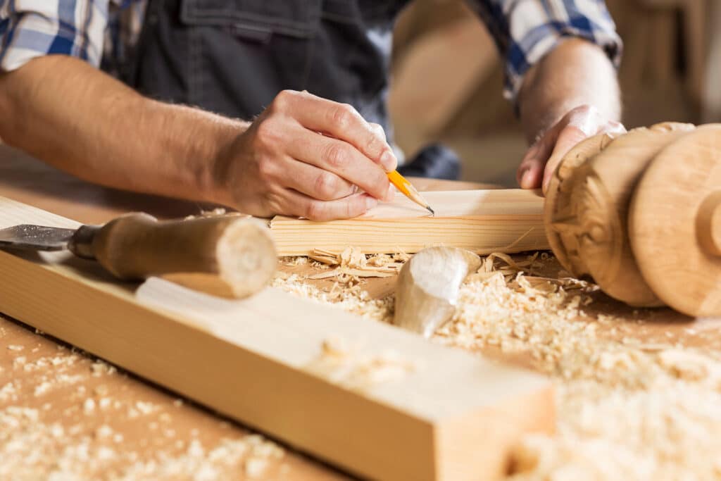 Menuiserie : marquage précis du bois avec un crayon Menuisier marquant des lignes sur une planche de bois avec un crayon, entouré de copeaux, près d'un tourné.