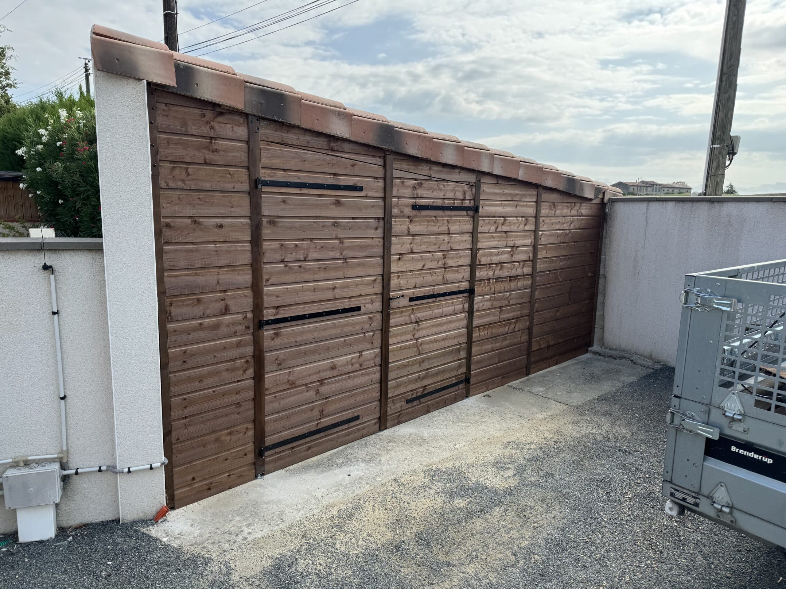 Abri de jardin en bois brun avec toit en tuiles rouges et noires, devant un mur blanc texturé et une remorque Brenderup.
